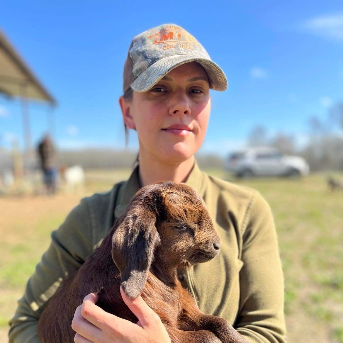 Woman holding a brown baby goat in a sunny outdoor setting, wearing a camouflage cap and a green jacket, with a blurred background of a farm and blue sky.